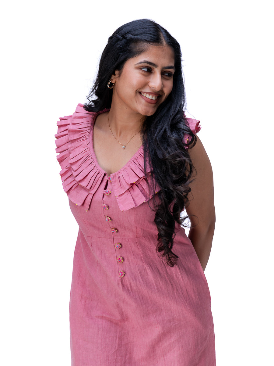 A woman with long, wavy black hair is wearing a dusty Ash pink sleeveless dress with a ruffled collar and front buttons, photographed against a solid white background.