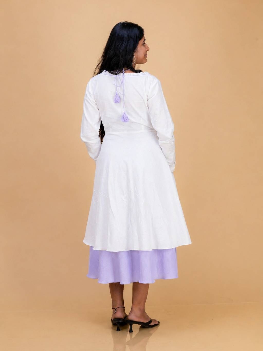 A woman is shown from the back wearing Ash pink & White long-sleeve tunic dress with a lavender skirt and black heels, set against a solid beige studio background.