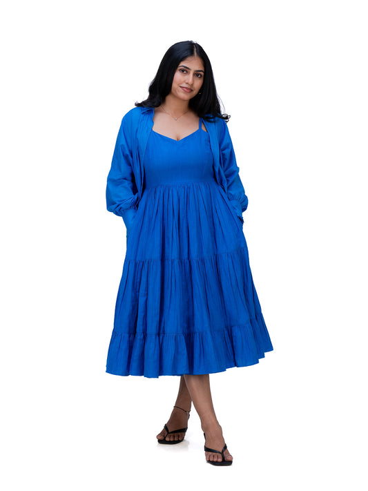 A woman modeling a bright royal Woad Blue  tiered midi dress with long sleeves and side pockets, paired with dark brown sandals, against a solid white background.
