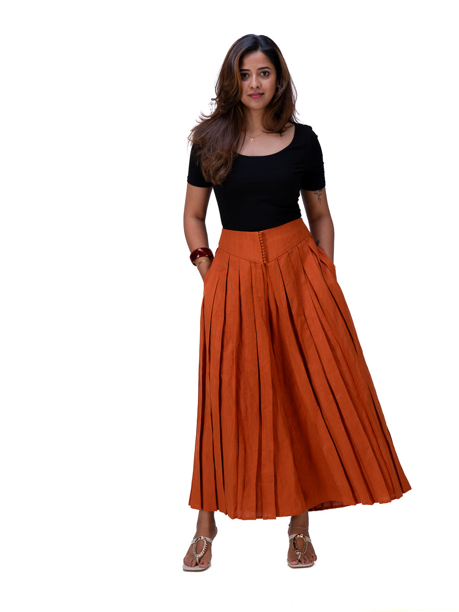 A woman modeling a black short-sleeve top and a high-waisted, rust-orange pleated maxi skirt with front button details, paired with Brazil Brown flat sandals, against a plain white background.