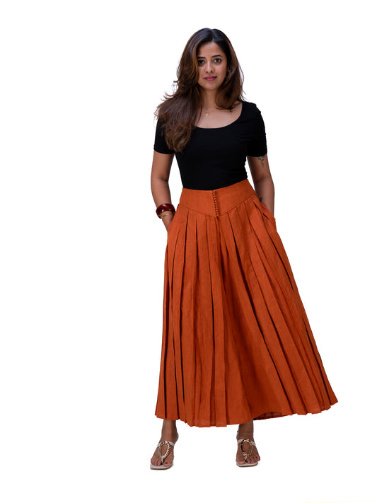 A woman modeling a black short-sleeve top and a high-waisted, rust-orange pleated maxi skirt with front button details, paired with Brazil Brown flat sandals, against a plain white background.