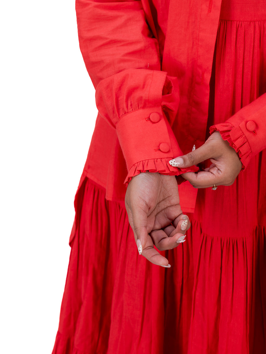 Close-up of a person wearing a bright Madder Red tiered dress with long ruffled sleeves and matching buttons, adjusting the cuff with both hands against a plain white background.
