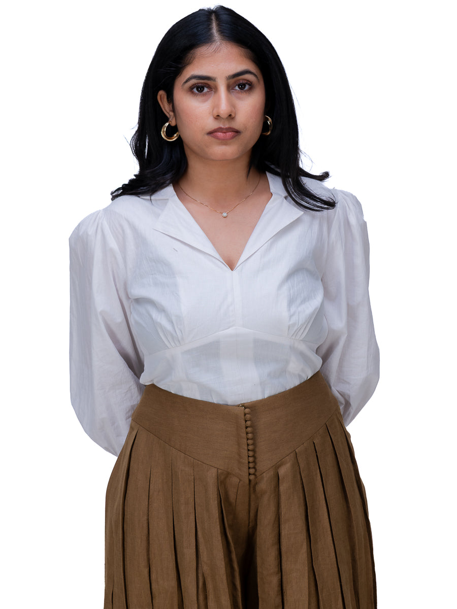 A woman wearing a white long-sleeve blouse with a V-neck and gathered waist, paired with a Oak Brown pleated skirt, against a plain white background.