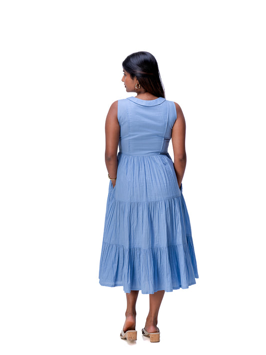 A woman viewed from the back wearing a sleeveless tiered midi dress in a dusty Nile Blue color, paired with tan platform sandals, against a plain white background.