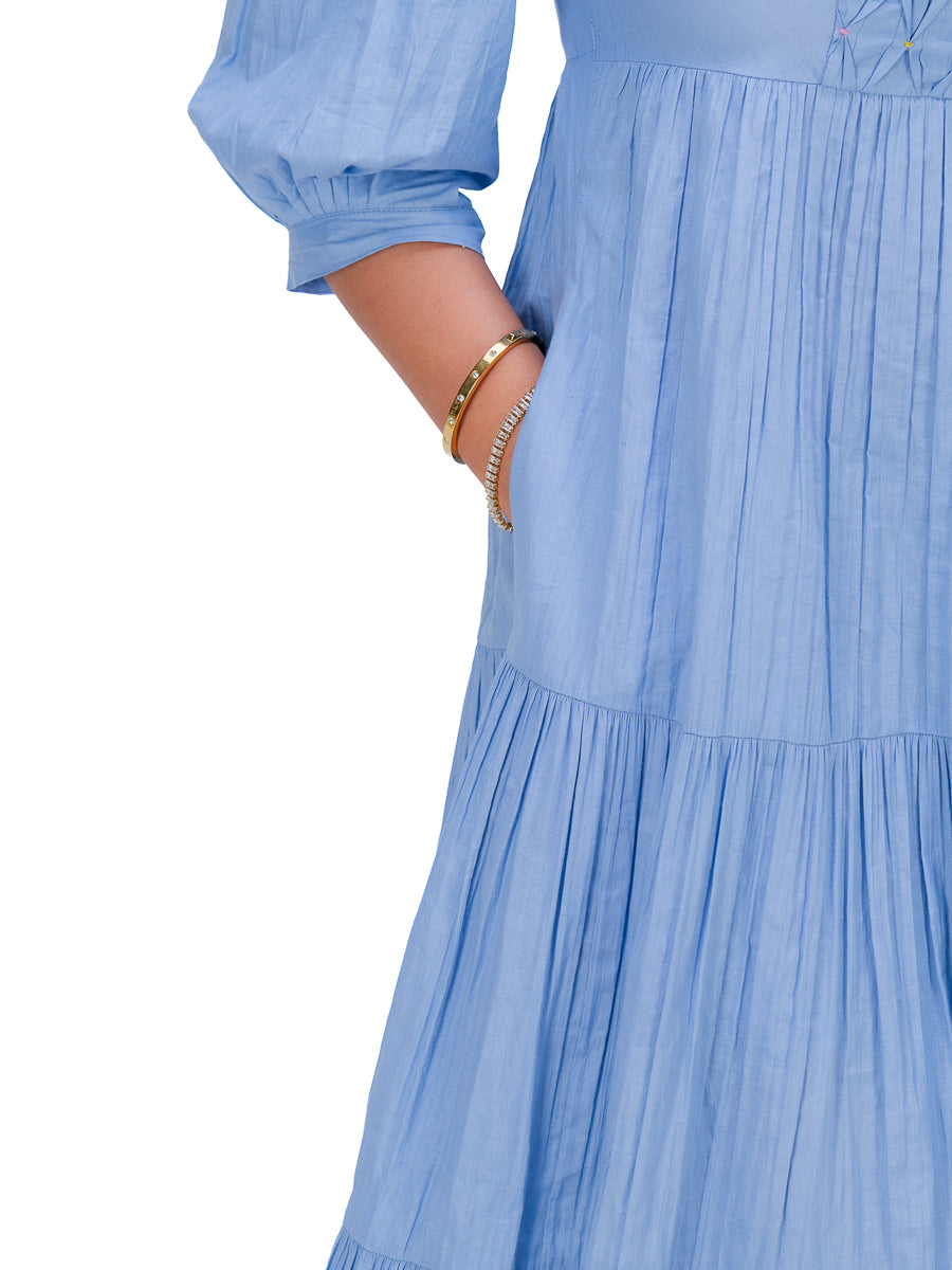 Close-up of a Nile Blue tiered dress with a pleated texture, featuring a sleeve with a gathered cuff and a side pocket containing gold-toned bracelets, shown against a plain white background.