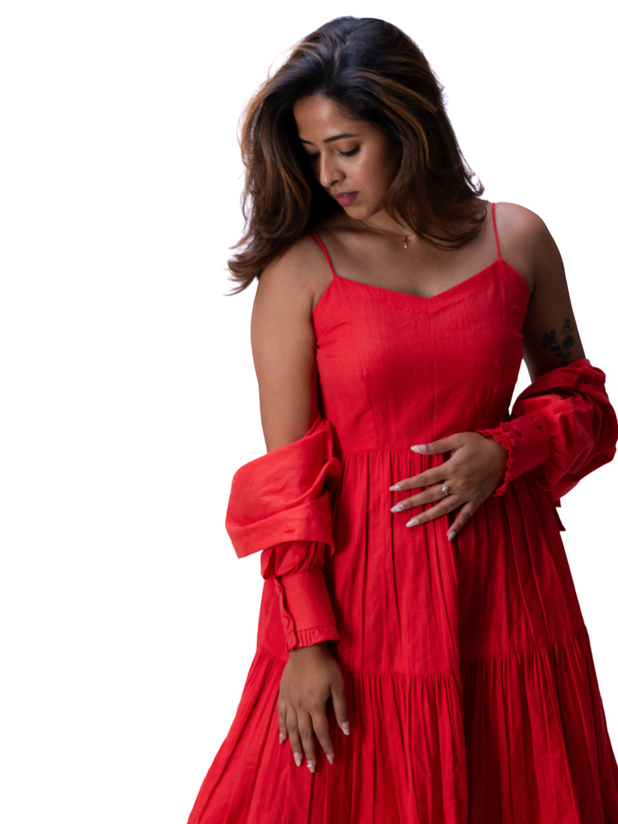 A woman wearing a bright Madder Red tiered maxi dress with thin spaghetti straps, a sweetheart neckline, and dramatic puffed long sleeves, posed against a solid white background.