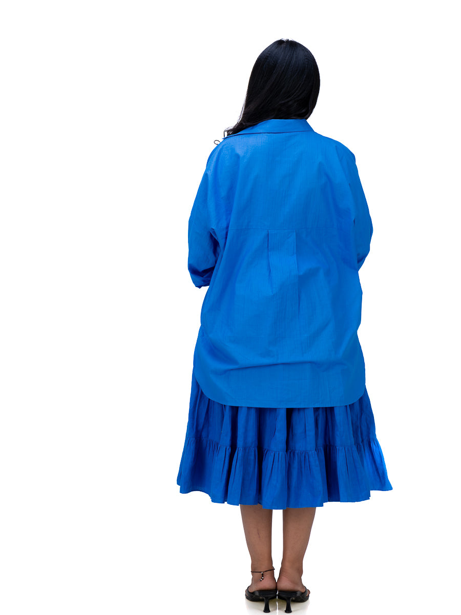 A woman shown from the back wearing a bright Woad Blue  long-sleeve collared shirt and matching blue tiered midi skirt, paired with black heels, against a plain white background.