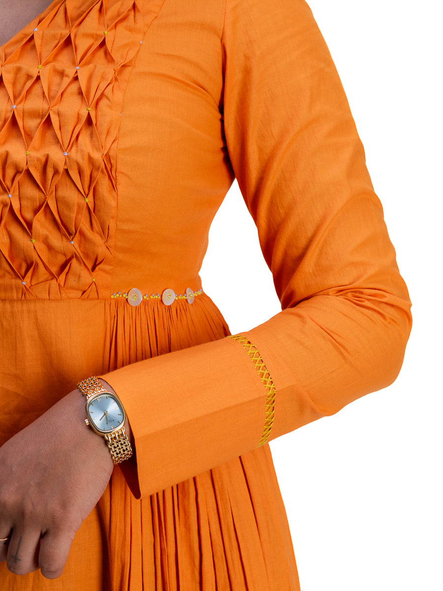 Close-up of a person wearing a bright Pomo orange long-sleeve dress with pleated detailing on the bodice, a thin decorative waistband, and gold embroidery on the cuff. A gold-tone watch is also visible on the wrist against a plain white background.