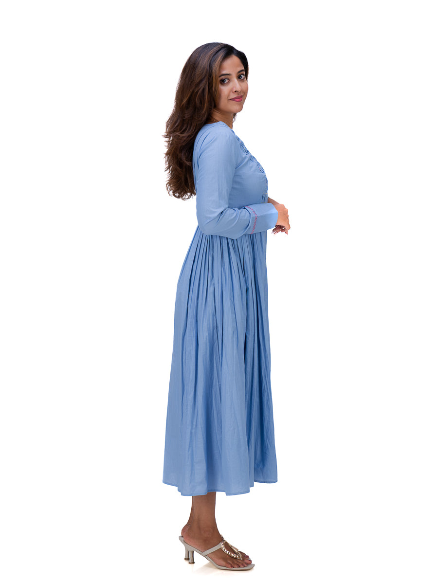 A woman modeling a light Nile Blue long-sleeve maxi dress with a gathered waist and pleated skirt, paired with brown heeled sandals, set against a plain white background.