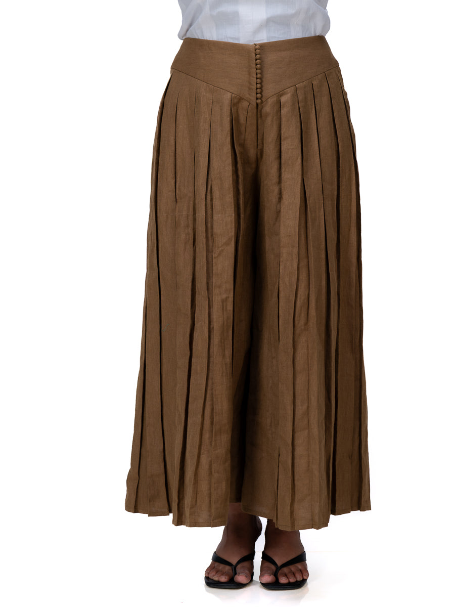 A person wearing a pair of wide-leg, pleated Oak Brown pants with a white top and black sandals, shown from the waist down against a plain white background.