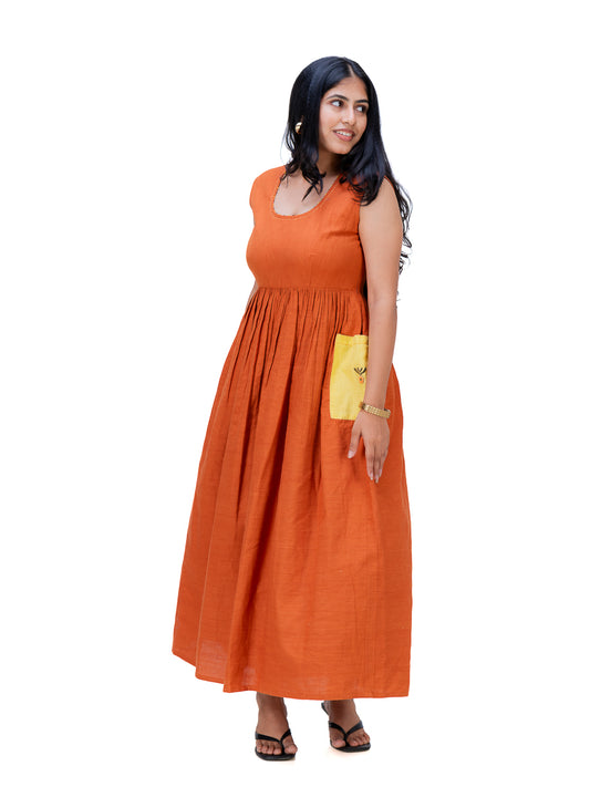 A woman modeling a sleeveless, scoop-neck maxi dress in a solid burnt Mud Orange color, featuring a gathered waist and a side pocket holding a yellow clutch, set against a plain white background.