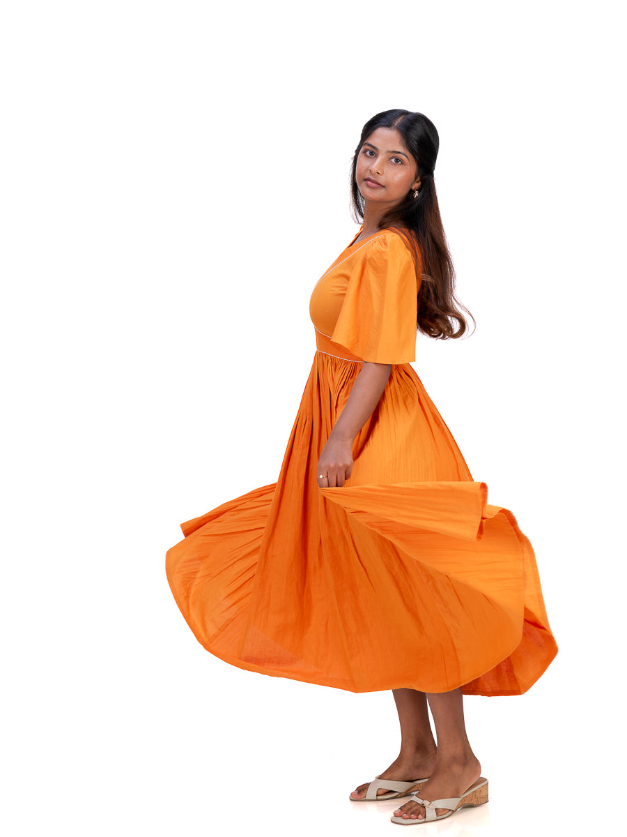 A woman wearing a bright Pomo orange short-sleeve midi dress with a flared skirt, captured from the side as she twirls to show off the fabric. She is wearing beige sandals against a solid white background.