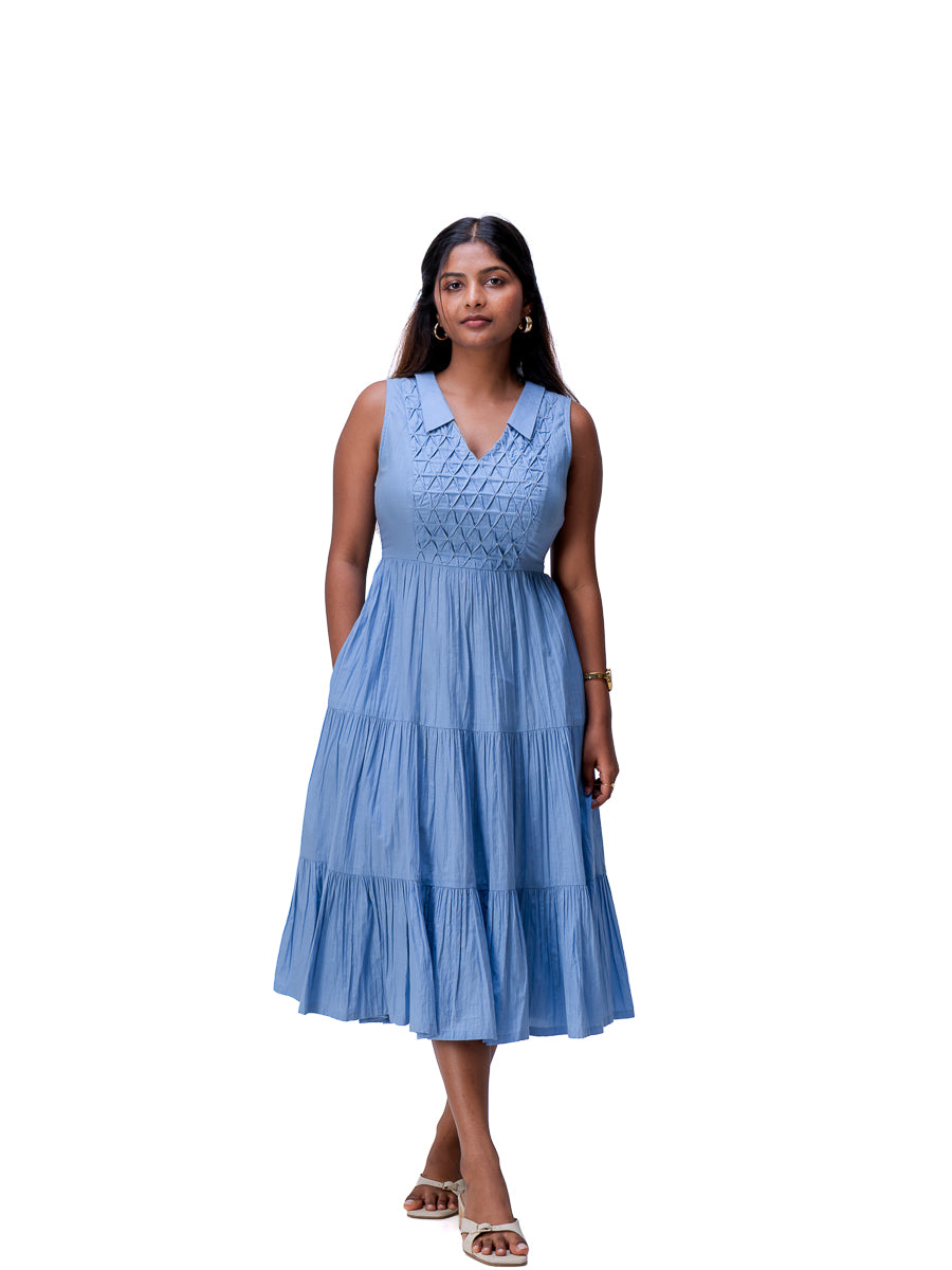 A woman modeling a sleeveless Nile Blue tiered midi dress with a V-neckline and embroidered chest detailing, set against a plain white background.
