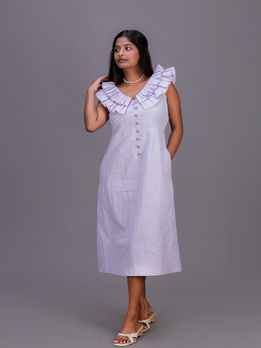 A woman modeling a sleeveless light Cherry Pink midi dress with a large ruffled collar and front button detail, standing against a solid gray studio background.