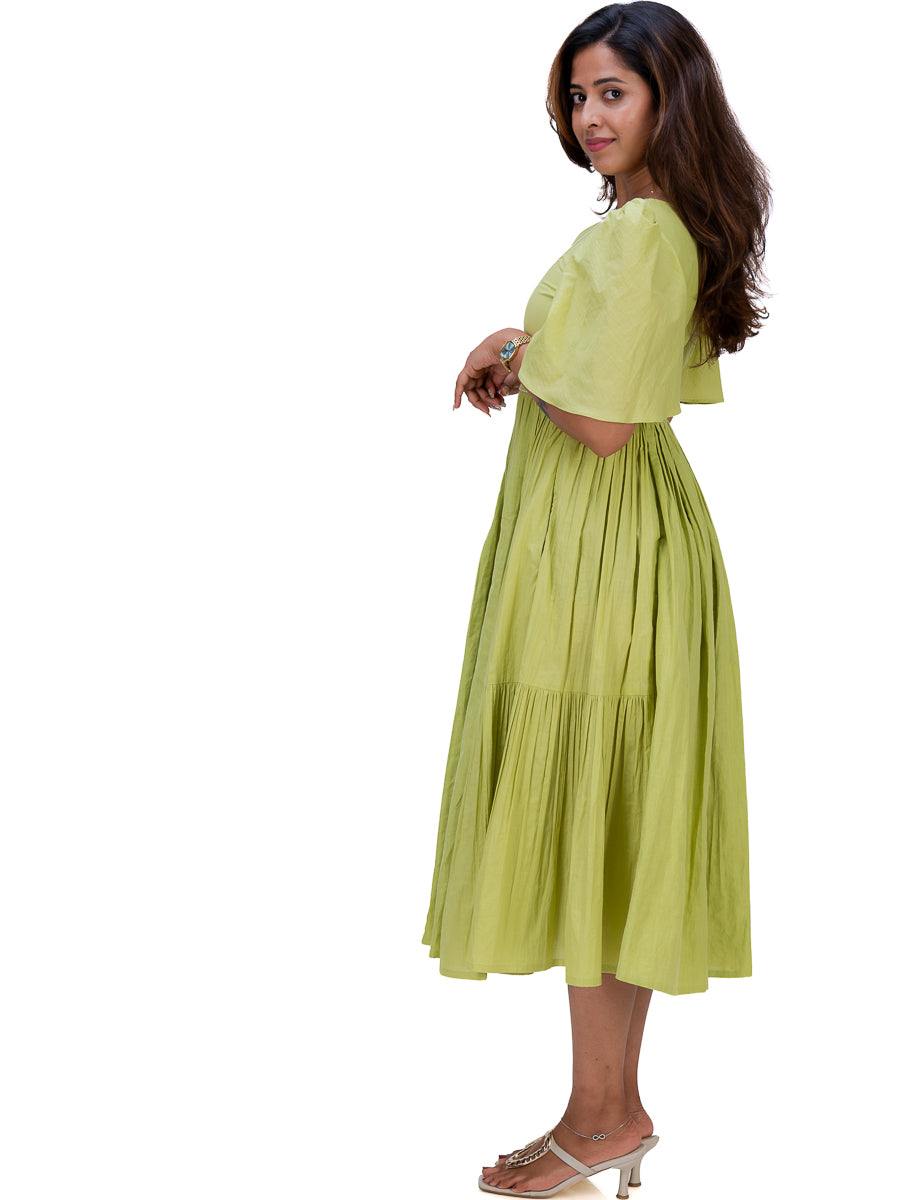 A woman modeling a light lime Dahlia Green tiered midi dress with short puff sleeves and side cutouts, paired with metallic heeled sandals, against a plain white background.