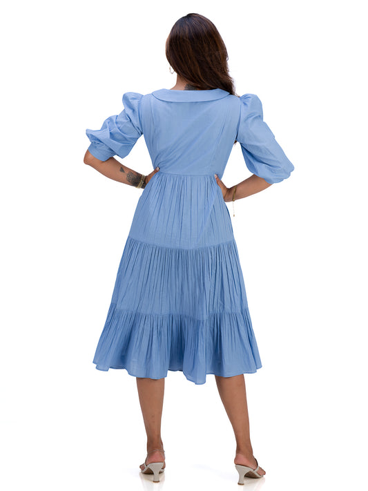 A woman viewed from the back wearing a Nile Blue midi dress with puffed short sleeves, a collared neckline, and a tiered skirt, paired with beige heeled sandals against a plain white background.
