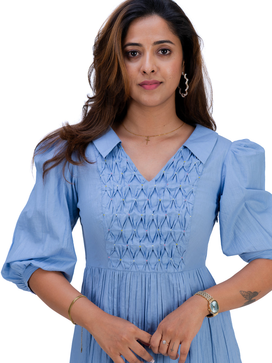 A woman wearing a Nile Blue dress with puff sleeves, a pointed collar, and a smocked bodice, shown against a plain white background.