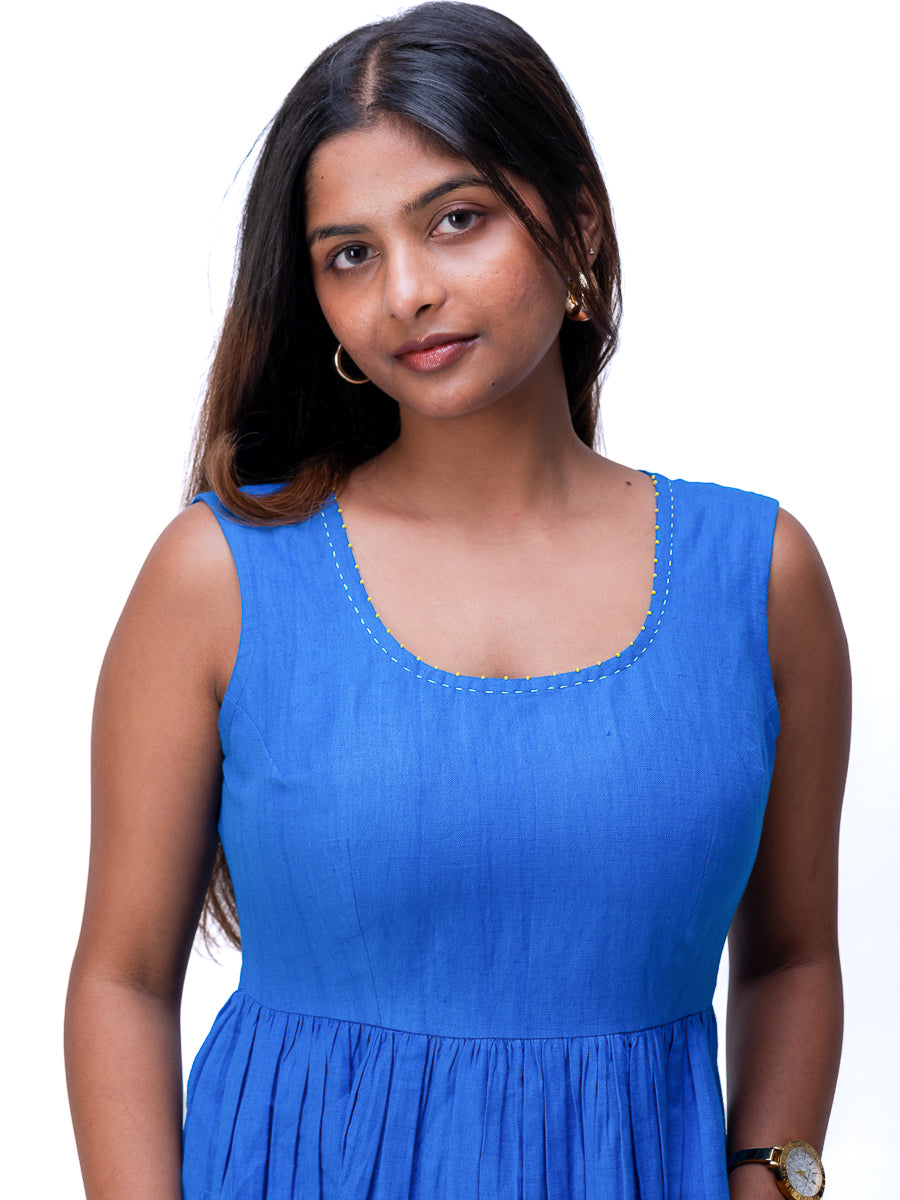 A woman wearing a sleeveless Woad Blue dress with a scoop neckline and subtle embroidered detailing around the neckline, photographed against a plain white background.