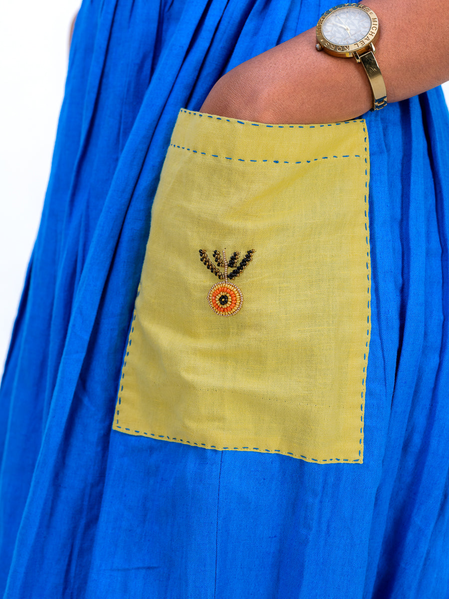 A close-up of a bright Woad Blue pleated garment featuring a large mustard yellow patch pocket with blue stitching and a small embroidered motif. A person's arm wearing a gold watch rests on the pocket.