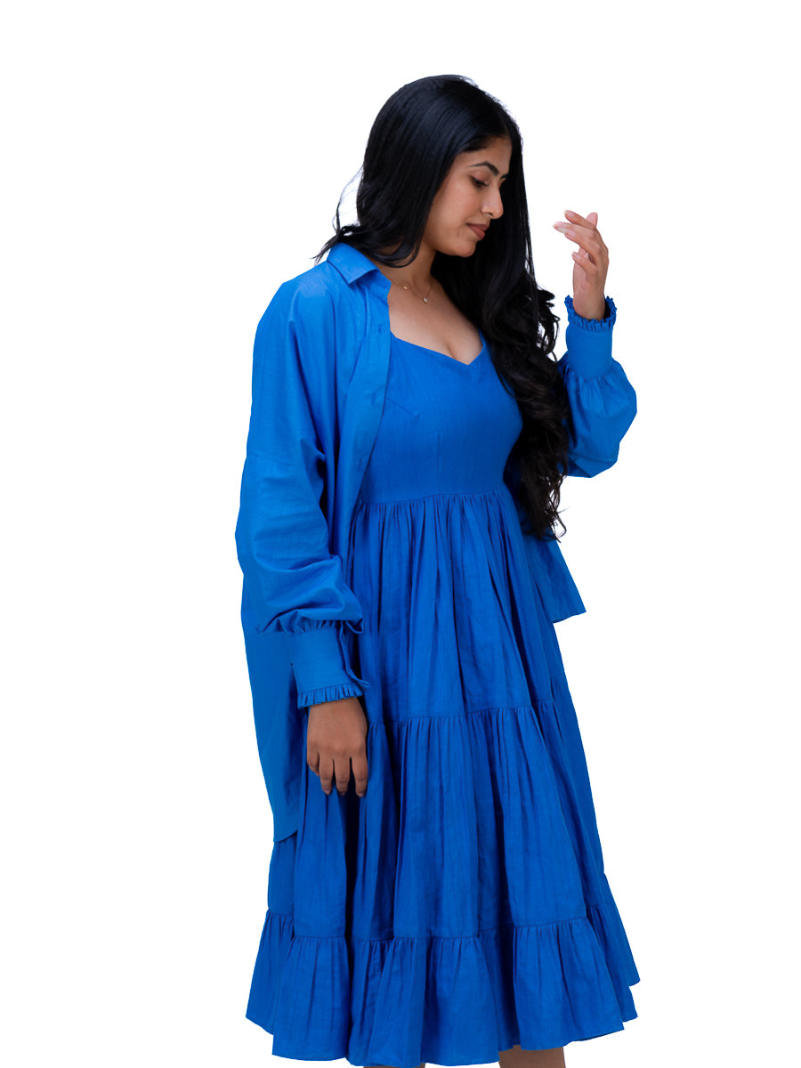 A woman modeling a bright royal Woad Blue  tiered midi dress with a matching long-sleeve button-up shirt jacket, set against a plain white background.