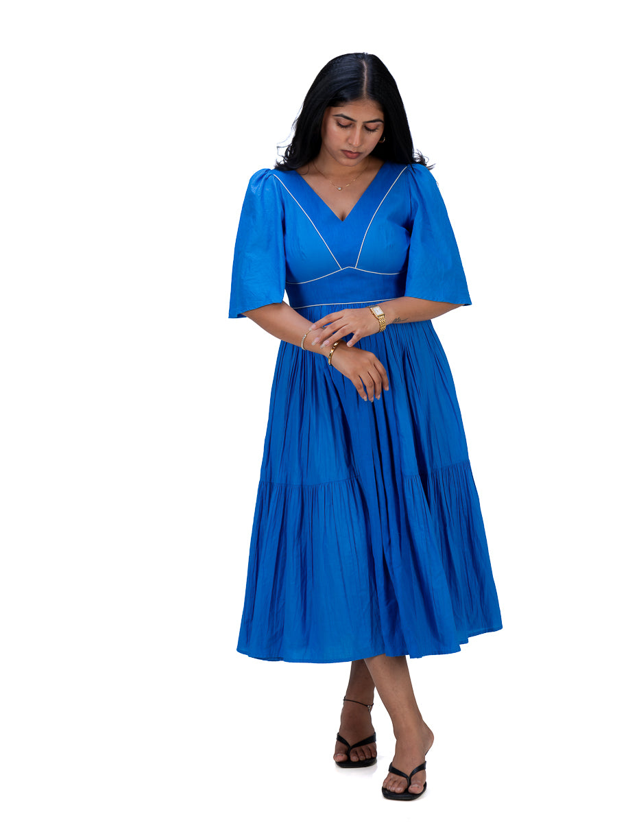 A woman modeling a bright Woad Blue midi dress with short flared sleeves, a V-neckline, and white contrast piping details, set against a plain white background.
