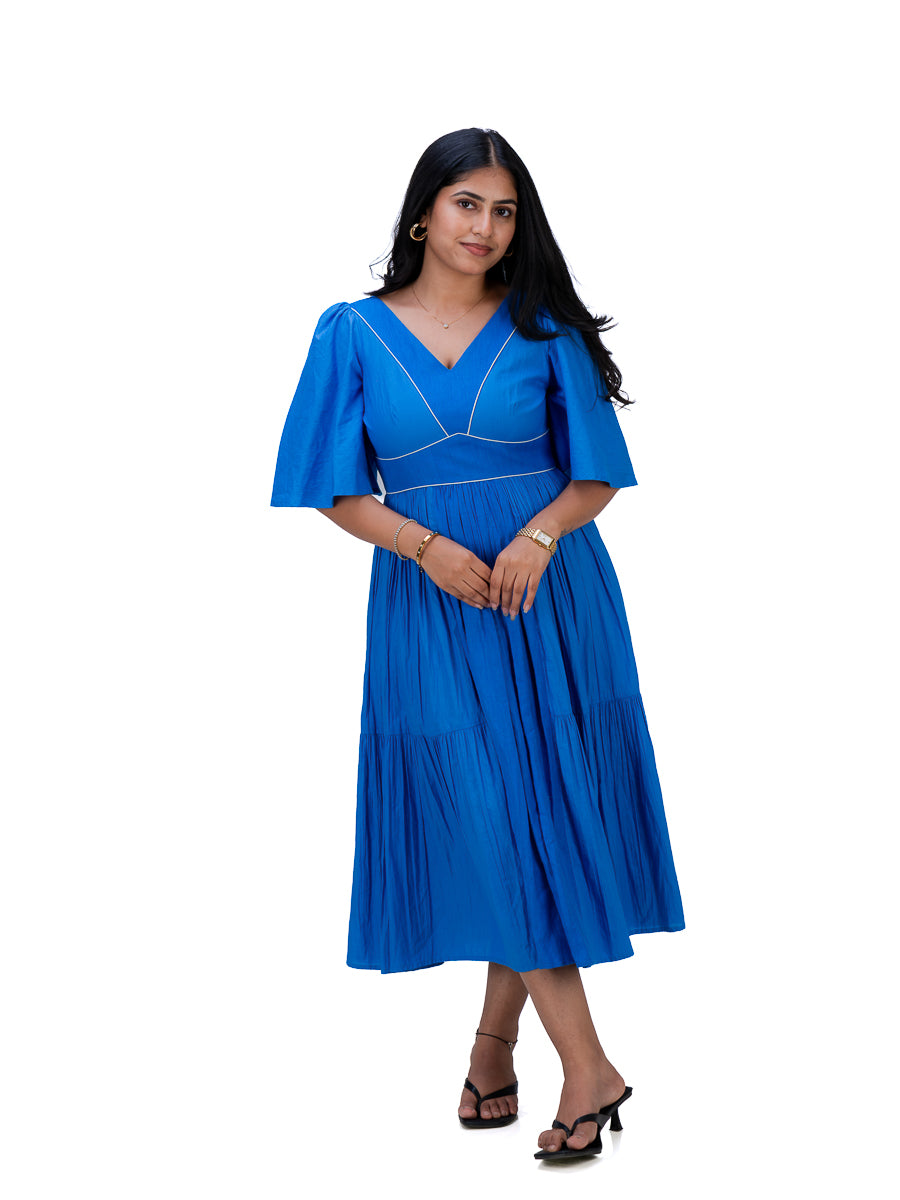 A woman modeling a bright Woad Blue midi dress with a V-neckline, short flutter sleeves, and a pleated tiered skirt, set against a plain white background.