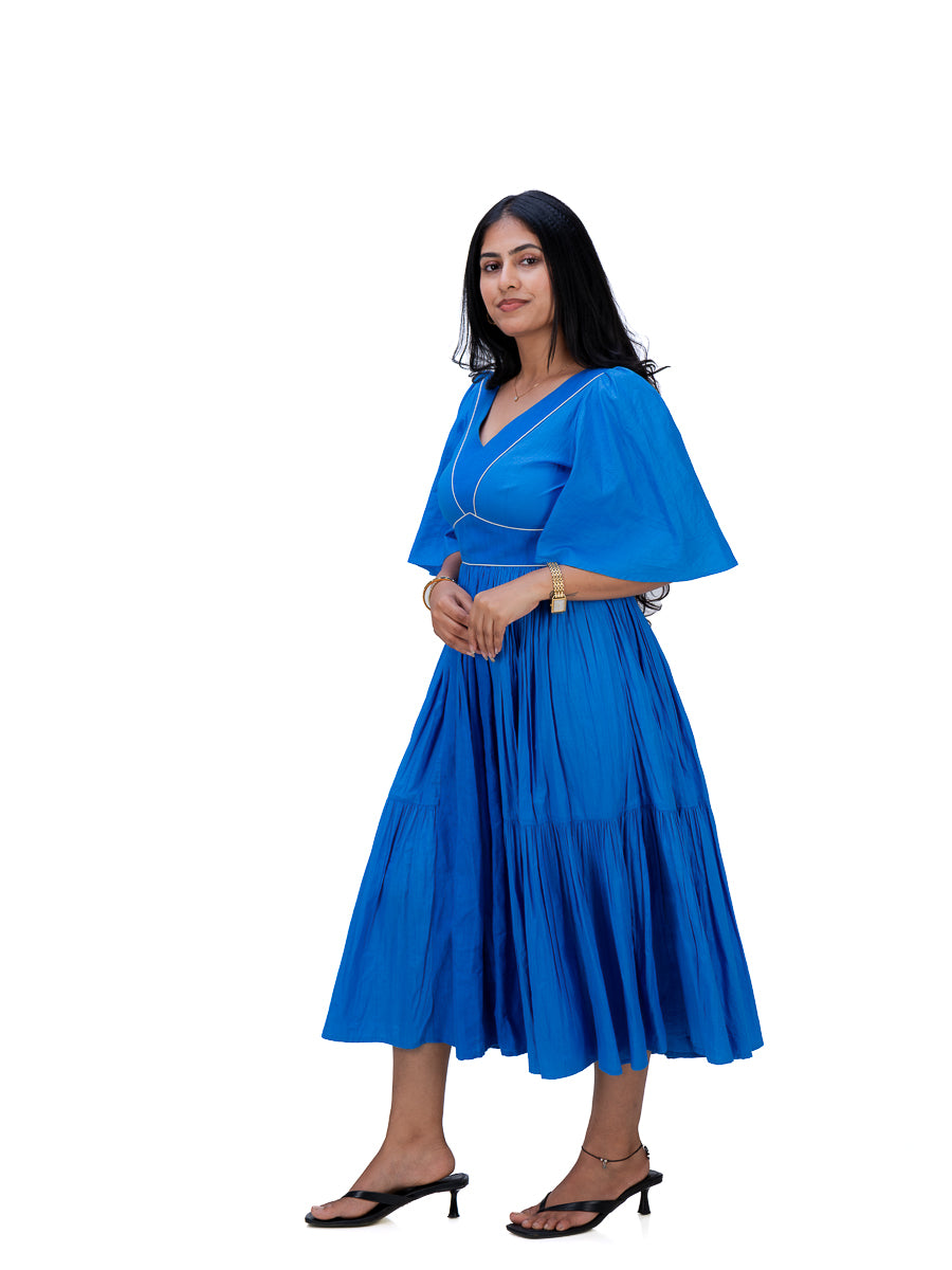 A woman modeling a bright Woad Blue midi dress with a V-neckline, flared sleeves, and a pleated tiered skirt, paired with black heeled sandals against a plain white background.