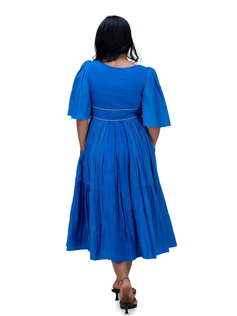 A woman viewed from the back wearing a bright royal Woad Blue midi dress with short puff sleeves, a fitted waist, and a pleated skirt, paired with dark heeled sandals against a plain white background.