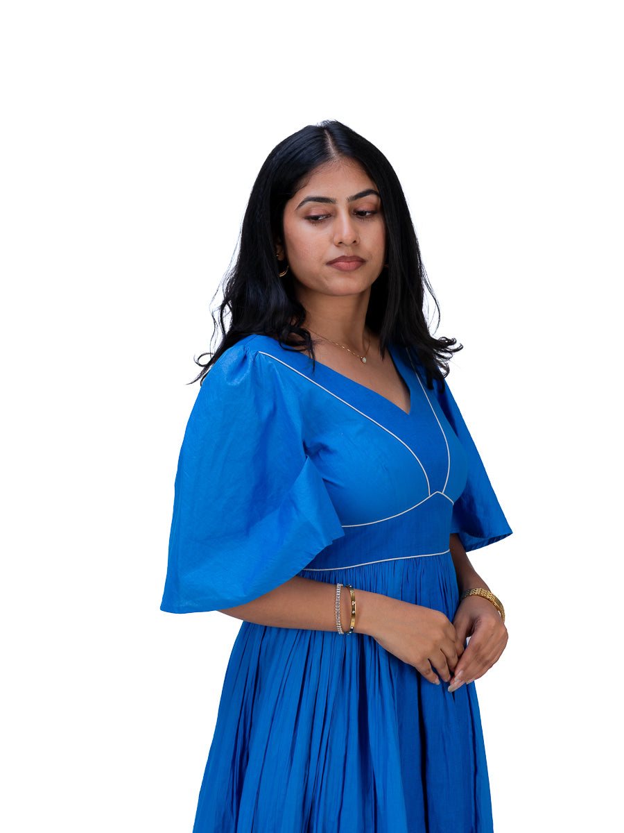 A woman wearing a bright Woad Blue dress with flutter sleeves and white contrast stitching, standing against a plain white background.