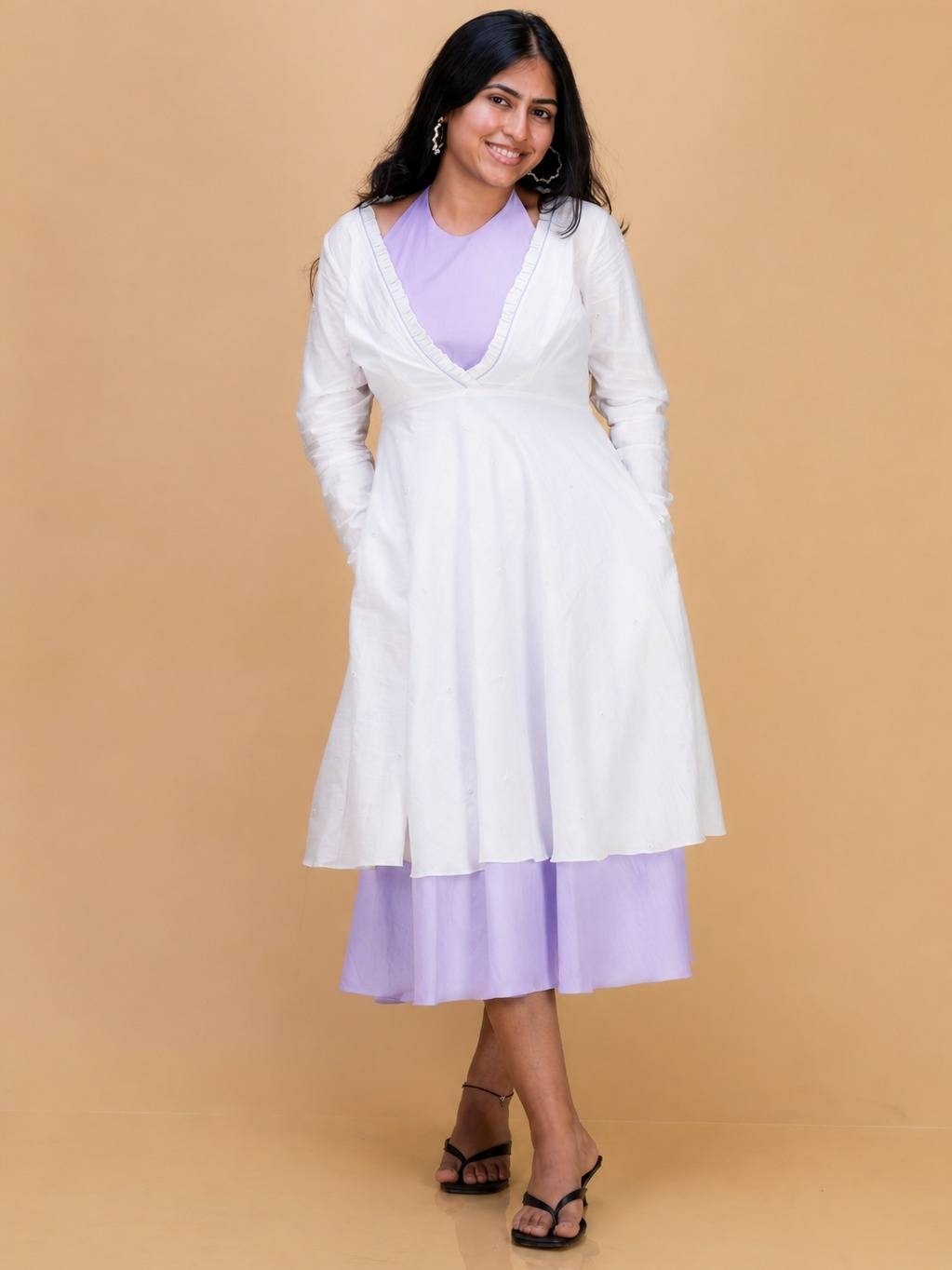 A woman modeling a white long-sleeve wrap-style dress over a Ash pink & White dress, standing against a solid beige studio background.