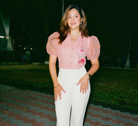 A woman posing outdoors at night wearing a light pink short-sleeve blouse with puff sleeves and a floral print, paired with white wide-leg trousers and brown ankle boots.