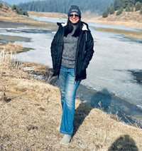 A person standing outdoors by a partially frozen river in a dry grassy landscape under a bright blue sky, wearing a black jacket, gray sweater, blue jeans, and a dark beanie.